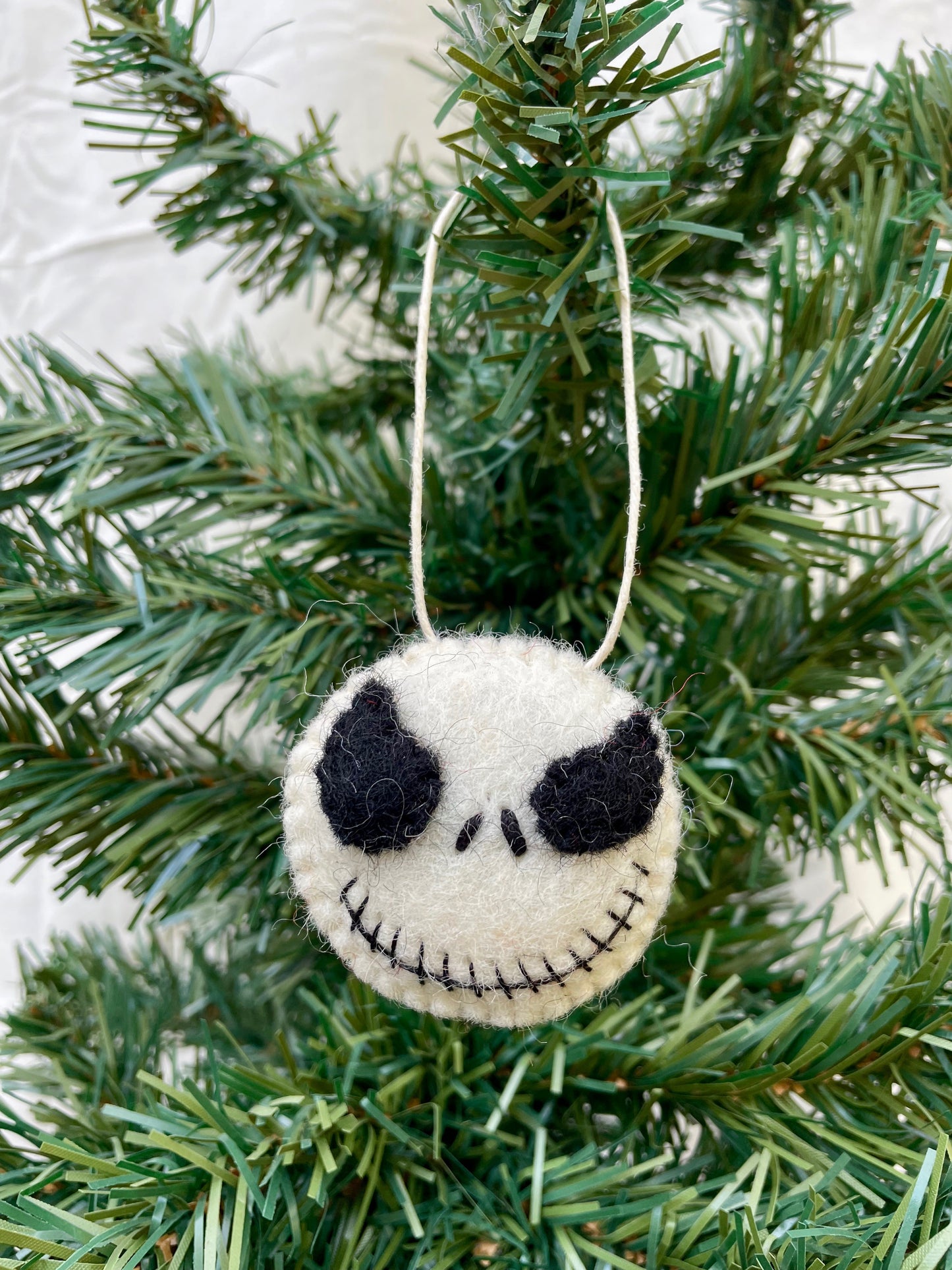 A handmade felt ornament in the shape of a skeleton's head, with black eyes and a grin, hanging from a green pine tree.