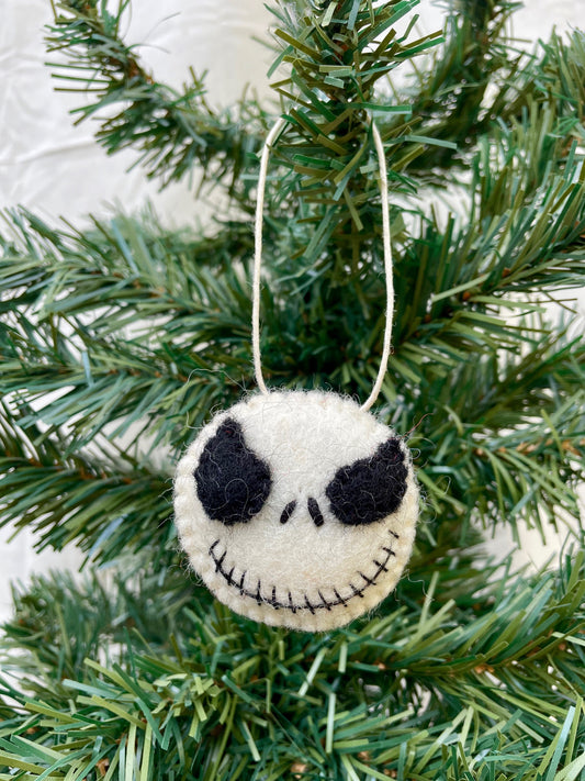 A handmade felt ornament in the shape of a skeleton's head, with black eyes and a grin, hanging from a green pine tree.